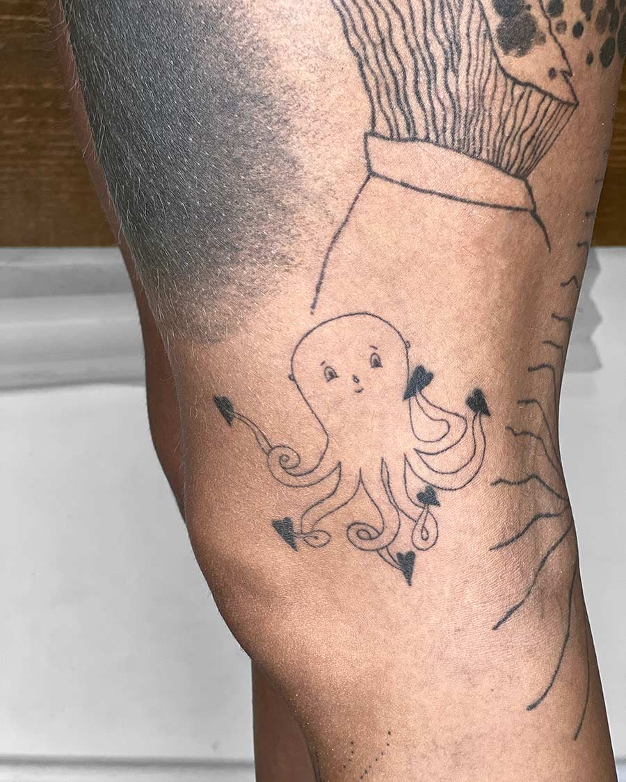 A minimalist tattoo of a smiling octopus with heart-shaped suckers on a person's leg. The tattoo is black ink and appears whimsical, with simple lines and a playful design. Other tattoo elements are visible nearby, suggesting a collection of body art. Done by tattoo artists in our fine line tattoo studio parlour shop Canggu, Bali