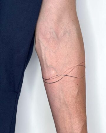 Close-up of a forearm with a minimalist armband tattoo. The fine line tattoo features three delicate, flowing black lines that wrap around the arm. This minimalist tattoo is a modern take on an armband. Done by tattoo artists in our fine line tattoo studio parlour shop Canggu, Bali