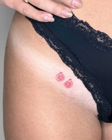 Close-up of a thigh with a minimalist red ink pomegranate tattoo. The delicate design features two halves of the fruit, a subtle and feminine pomegranate tattoo. Black lace underwear frames the skin, adding a touch of elegance. Done by tattoo artists in our fine line tattoo studio parlour shop Canggu, Bali