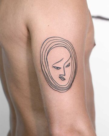 A minimalist tattoo design of a stylized face on a person's upper arm. The black ink tattoo features a simple outline of a face within a series of concentric circles. This tattoo art example showcases a minimalist tattoo style.