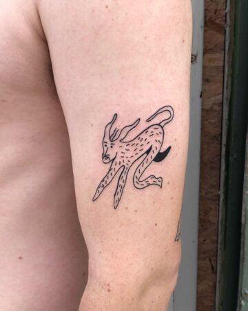 A minimalist tattoo of a creature with a human-like face and animal body, inked in black on a person's arm. The design features elongated limbs and a curved tail, showcasing a unique tattoo design.
