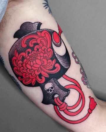 A vibrant neo-traditional tattoo on an arm features a black and red daruma doll adorned with a large red chrysanthemum. A tiny skull is visible on the doll's base, and red cords hang down. Japanese (Irezumi/Wabori) tattoo studio Canggu Bali