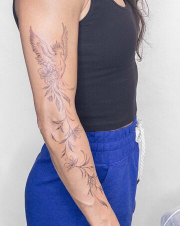 A woman's arm with an elegant phoenix tattoo flowing from shoulder to forearm, intertwined with delicate floral designs. She wears a black tank top and blue sweatpants, standing against a white wall.
