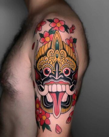 Bold tattoo of a Balinese Barong mask with a protruding tongue, surrounded by vibrant red and pink cherry blossoms on a man's upper arm. Japanese (Irezumi/Wabori) tattoo studio Canggu Bali