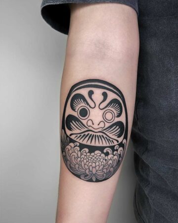 Close-up of a bold, black ink Daruma doll tattoo on a person's arm. The traditional Japanese doll design includes stylized facial features and a chrysanthemum flower pattern at the base. Japanese (Irezumi/Wabori) tattoo studio Canggu Bali