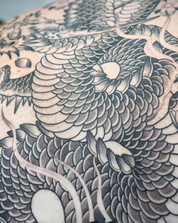 Close-up of a detailed black and gray dragon tattoo covering a large area of skin, showcasing intricate scale work and traditional Japanese tattoo art.