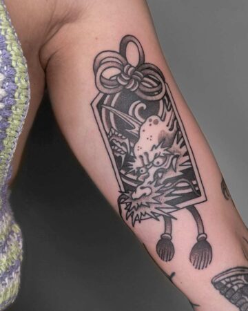 Close-up of a traditional black and gray tattoo of a Japanese dragon head framed in a rectangular charm with a decorative knot at the top and tassels hanging below, inked on a person's arm. Japanese (Irezumi/Wabori) tattoo artist studio Canggu Bali