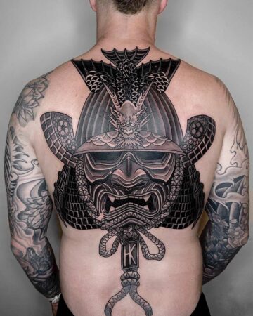 Detailed back tattoo of a samurai mask and helmet. The black and grey ink depicts intricate armor plating, a menacing face with sharp teeth, and a dragon crest on top. Japanese (Irezumi/Wabori) tattoo artist studio Canggu Bali