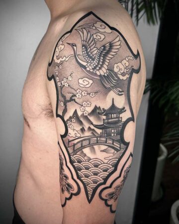 Detailed black and gray tattoo on an arm depicting a crane in flight above a pagoda, mountains, and bridge within an ornate frame. Japanese Oriental Tattoo made in Bali