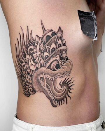 Detailed black ink tattoo of a Balinese Barong mask on a woman's ribcage. The intricate design features floral elements and traditional headdress. Japanese (Irezumi/Wabori) tattoo artist studio Canggu Bali