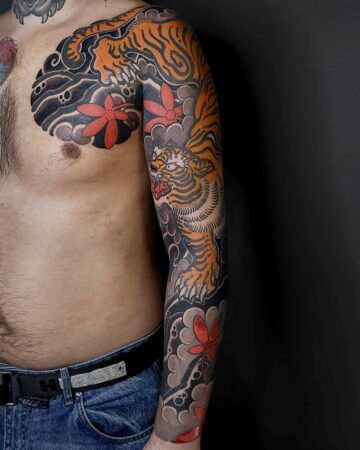 Torso and arm featuring a vibrant Japanese tiger tattoo. The traditional Irezumi design includes orange tiger stripes, stylized clouds, and red floral accents against a black background, extending from the chest to the forearm. Japanese (Irezumi/Wabori) tattoo artist studio Canggu Bali