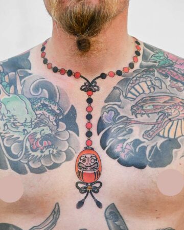Torso with vibrant Japanese tattoos, including a Daruma doll necklace tattoo in red and black. Chest also features dragon and other traditional imagery.