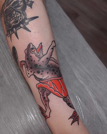 Traditional tattoo of a frog wearing a red skirt, seemingly dancing with its mouth open. Black spots cover its body, and another tattoo is partially visible above it. Japanese (Irezumi/Wabori) tattoo artist studio Canggu Bali