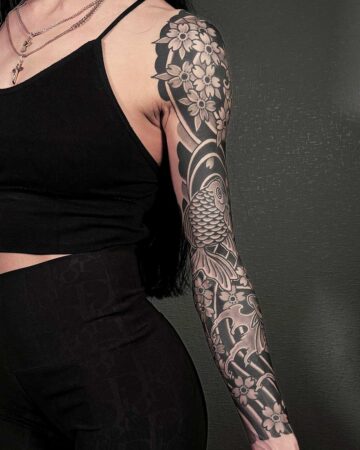 Woman's arm with a striking black and gray Japanese-style tattoo sleeve featuring a koi fish, cherry blossoms, and wave patterns. The tattoo extends from her shoulder to her wrist. Japanese (Irezumi/Wabori) tattoo artist studio Canggu Bali