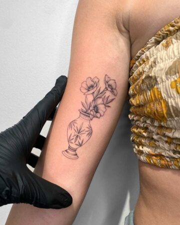 A delicate floral tattoo gallery image shows a vase etched with stars, filled with three simple flowers on the inner arm. A black-gloved hand gently holds the arm to display the artwork.