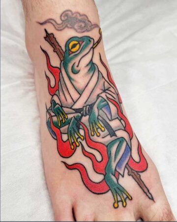 Vibrant color traditional tattoo of a frog in a gi, smoking a pipe with a branch and surrounded by red flames and a cloud. Bali tattoo studio.