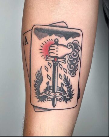 A bold color traditional tattoo on an arm features a stylized ace playing card with a powerful fist holding a sword, surrounded by a laurel wreath and dramatic clouds. This tattoo showcases vibrant color and classic tattoo art, created in a Canggu Bali tattoo studio.