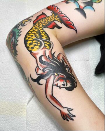 Vibrant color traditional tattoo of a mermaid with a yellow and red scaled tail, black hair, and red accents on an arm. Bali tattoo studio. Tattoo artist in Canggu Bali.