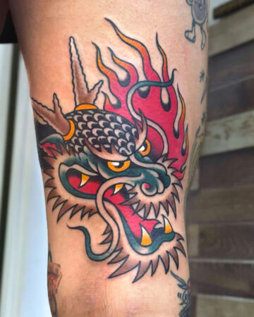 Vibrant color traditional tattoo of a fierce dragon head with red flames, sharp teeth, and yellow eyes, showcasing bold lines and a classic American traditional tattoo style. Bali tattoo studio.