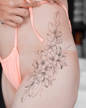 A woman's hip and thigh are adorned with a delicate floral tattoo featuring outlined cherry blossoms and shaded leaves, complemented by a peach-colored bikini bottom.