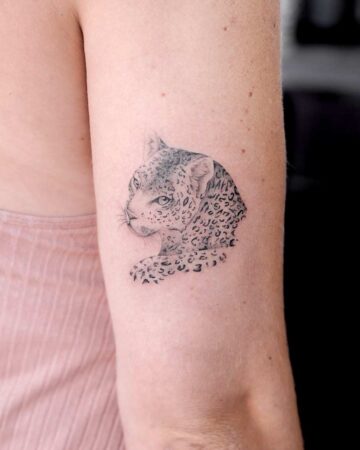 Close-up of a delicate leopard tattoo on a woman's upper arm. The black ink tattoo features fine line work and detailed shading, showcasing the big cat's face and shoulder. A striking lion tattoo alternative.
