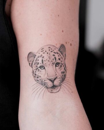 Close-up of a delicate leopard tattoo on an arm. The black ink tattoo features the leopard's face with detailed spots and whiskers, showcasing a popular lion tattoo design.