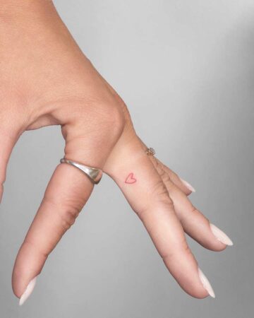 Close-up of a hand with a delicate red heart finger tattoo on the side of the ring finger. A silver ring is worn on the pinky finger. Part of a fingers tattoo gallery.