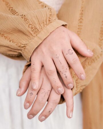 Close-up of hands with delicate finger tattoos: celestial designs including a crescent moon, stars, and constellations. Beige top and neutral nail polish complement the minimalist aesthetic. Fingers tattoo gallery.