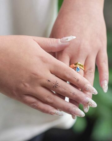 Woman's hands displaying delicate finger tattoos and almond-shaped nails with floral designs, showcasing a unique style from a fingers tattoo gallery. A gold ring with a blue gemstone adorns her finger.