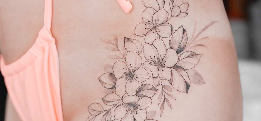 A woman's hip and thigh are adorned with a delicate floral tattoo featuring outlined cherry blossoms and shaded leaves, complemented by a peach-colored bikini bottom.