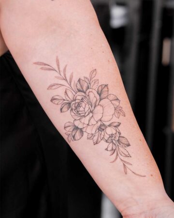 A delicate floral tattoo featuring detailed roses peony and leaves graces a person's forearm. The fine line work creates an elegant and botanical design.