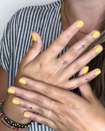Woman's hands with yellow nail polish and delicate finger tattoos, including a crescent moon and tiny stars. A stylish fingers tattoo gallery image. Custom design from our Canggu tattoo artist in Bali.
