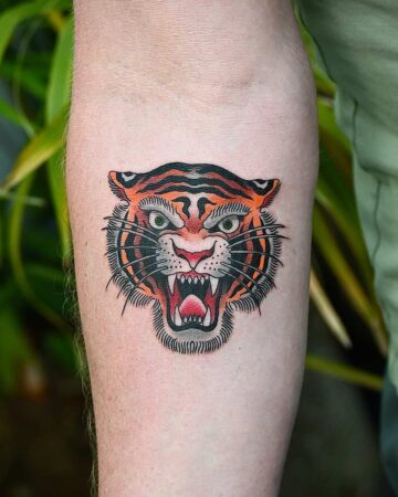 Close-up of an arm tattoo featuring a traditional-style snarling tiger head with orange stripes, green eyes, and a red open mouth showing teeth. A classic lion tattoo design.