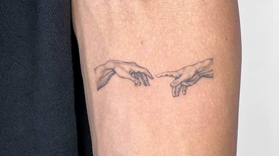 Close-up of a forearm featuring a Micro Realism Tattoo of Michelangelo's "The Creation of Adam," accompanied by the French text "l'art sauve des vies" (art saves lives).