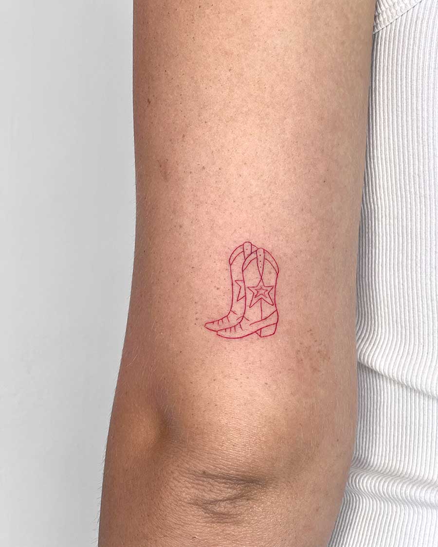 Minimalist red line art tattoo of cowboy boots with a star detail on a person's upper arm. Created in the "FLT Tattoo" a multi style tattoo studio in Canggu, Bali, for your Bali tattoo needs.