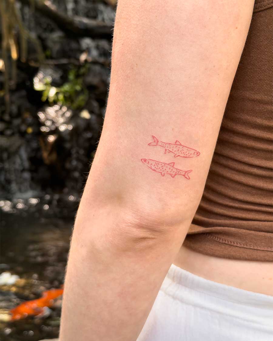 Two small, minimalist red fish tattoos are shown on a person's upper arm. Created in the "FLT Tattoo" a multi style tattoo studio in Canggu, Bali, this red tattoo is a unique Bali tattoo.
