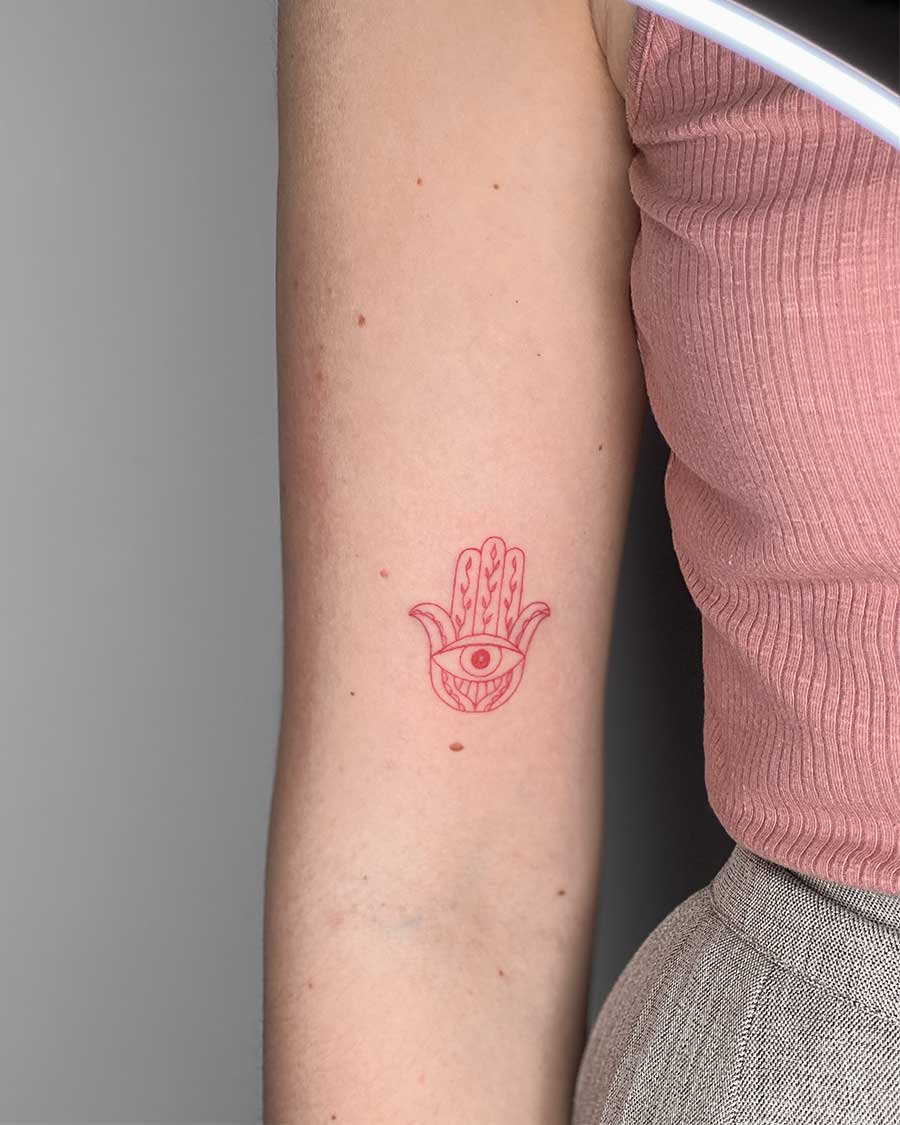 Close-up of a small, red hamsa hand tattoo with an eye in the palm, inked on the inner forearm of a person. Created in the FLT Tattoo, a multi-style tattoo studio in Canggu, Bali, known for Bali tattoos.