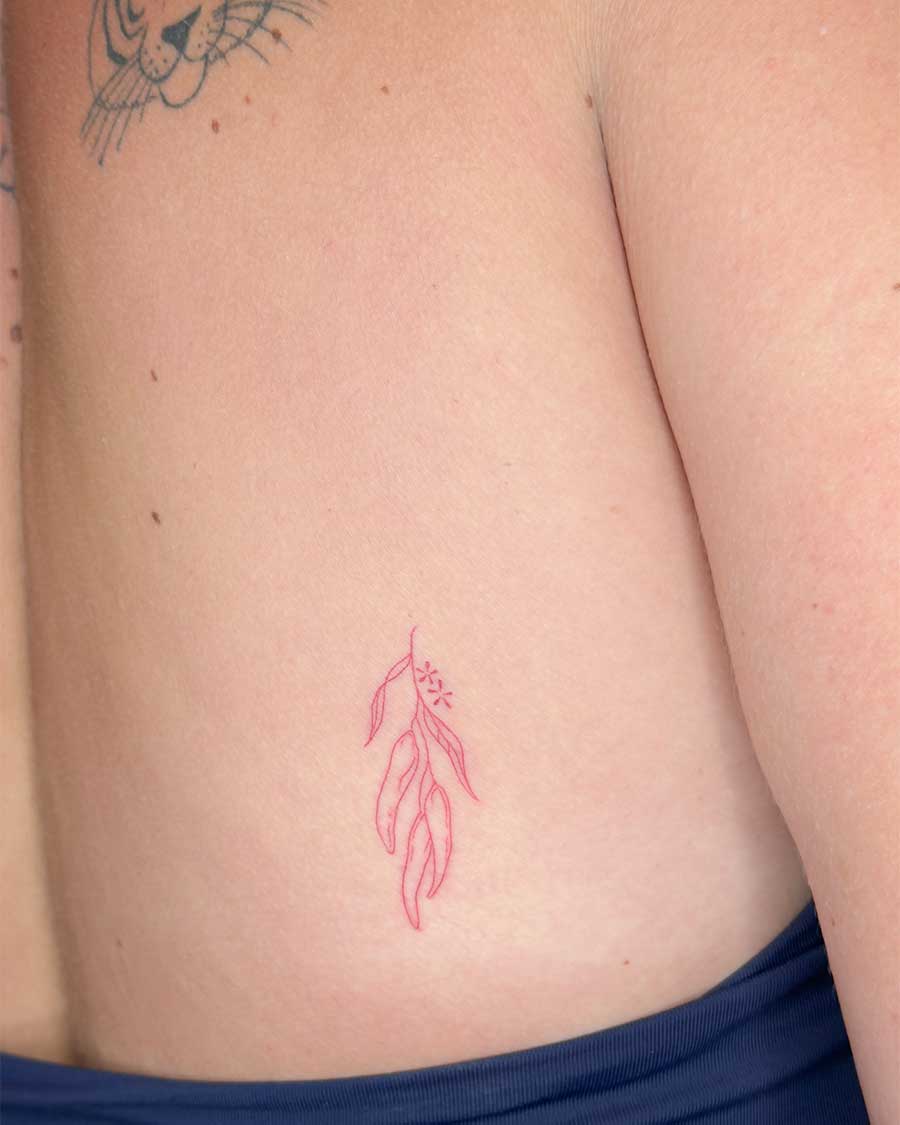 A delicate red ink tattoo of a eucalyptus branch with leaves and seed pods is inked on the side of a person's torso. Created in the FLT Tattoo, a red tattoo studio in Canggu, Bali.