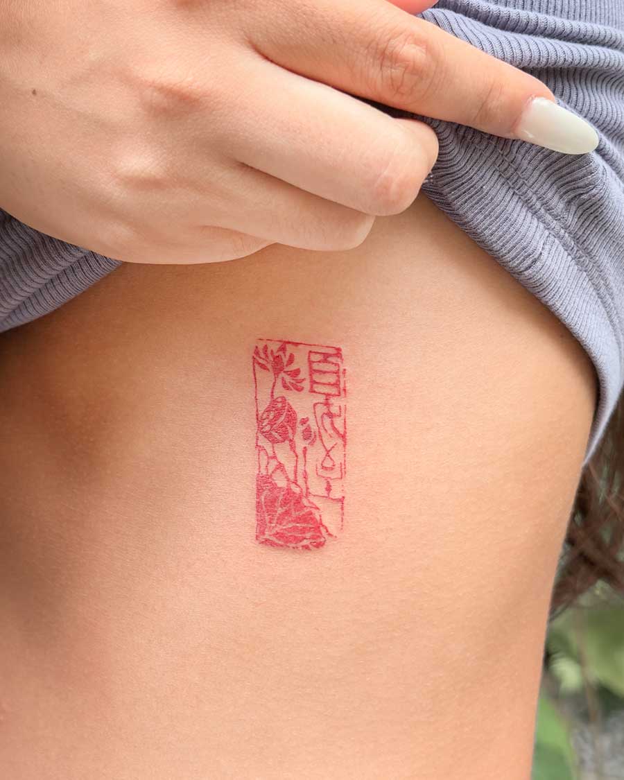 A vibrant red tattoo resembling an ancient seal is inked on a person's ribcage, showcasing intricate floral and symbolic designs within its rectangular frame. Created in the FLT Tattoo, a multi-style tattoo studio in Canggu, Bali, offering unique Bali tattoo experiences.