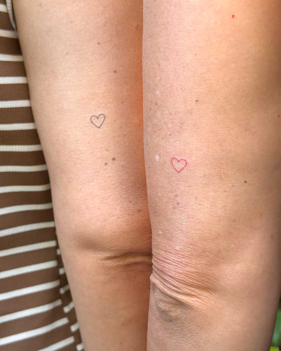 Close-up of two small, minimalist heart tattoos on fair skin. One red heart tattoo is on the right knee, and a black heart tattoo is on the left knee. Created in the "FLT Tattoo" a multi-style tattoo studio in Canggu, Bali. Bali tattoo studio, FLT Tattoo Canggu.