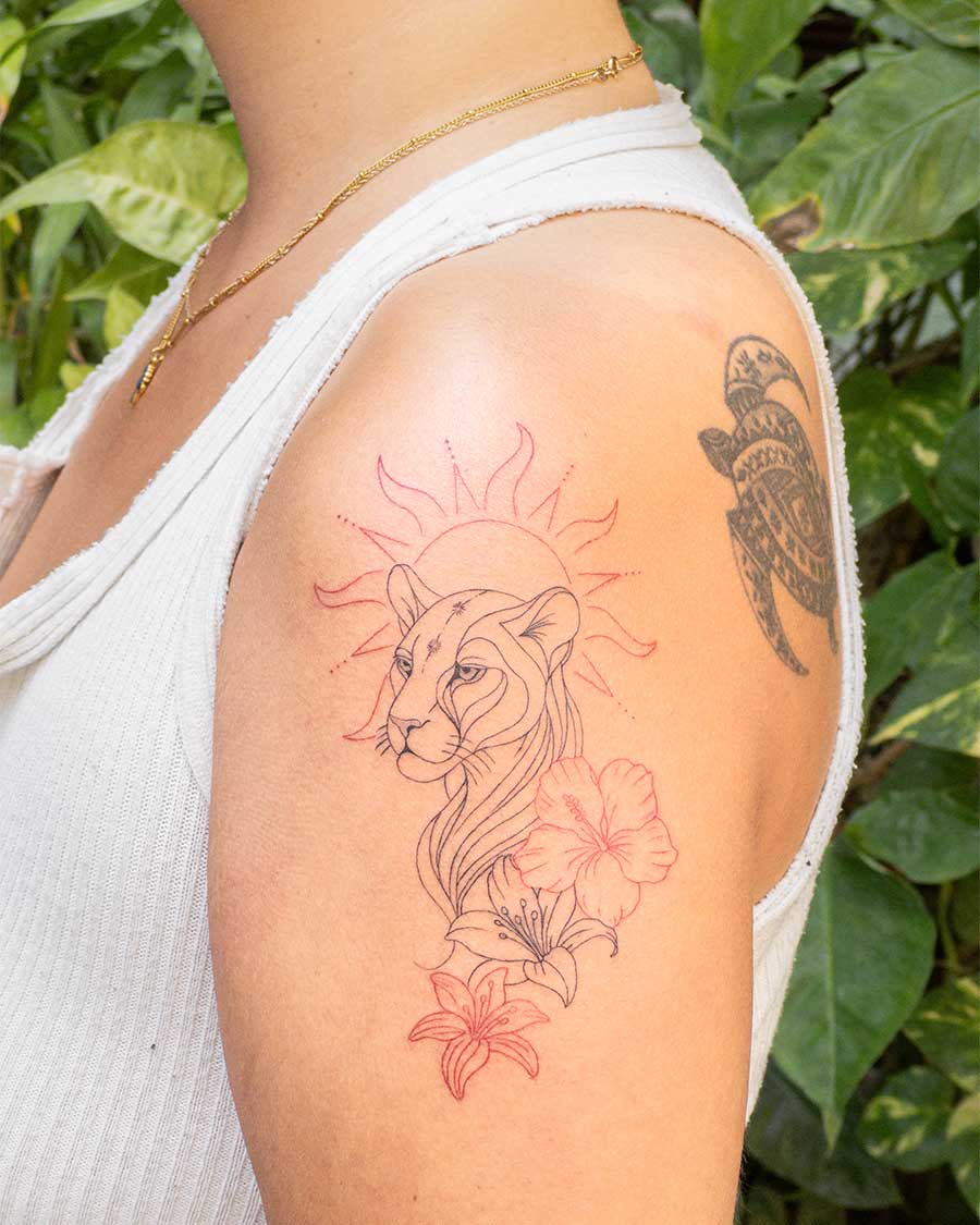 A red ink tattoo on a woman's upper arm features a stylized lioness profile with a sunburst behind it, accompanied by delicate hibiscus and lily flowers. Created in the FLT Tattoo studio, a top Bali tattoo destination for red ink and unique designs.