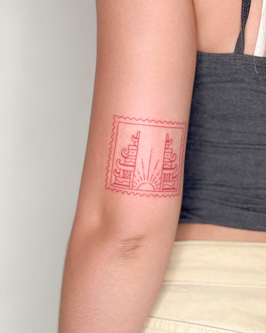 A minimalist red tattoo of a stylized temple gate with a sunrise peeking through, resembling a postage stamp, inked on an arm. Created in the FLT Tattoo, a multi-style tattoo studio in Canggu, Bali, offering unique Bali tattoo experiences.