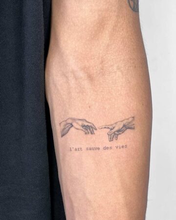 Close-up of a forearm featuring a Micro Realism Tattoo of Michelangelo's "The Creation of Adam," accompanied by the French text "l'art sauve des vies" (art saves lives).