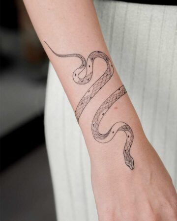 Elegant snake tattoo wraps around a fair-skinned wrist. Delicate black ink outlines the snake's form and subtle spotted pattern, showcasing a minimalist snake tattoo design. Created by our tattoo artists in Canggu, Bali