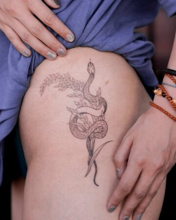 Detailed line art tattoo on a woman's side depicting a snake entwined with leafy branches. Her hand gently holds up a blue shirt, revealing the delicate tattoo design.Created by our tattoo artists in Canggu, Bali