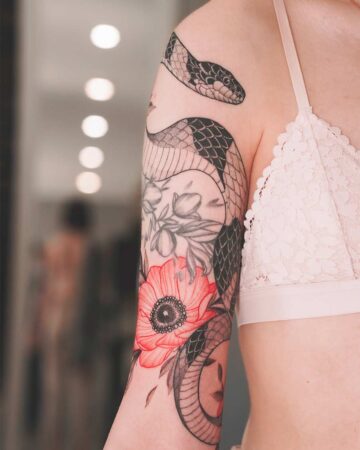 Woman's arm with a snake tattoo intertwined with red poppy flowers. The tattoo covers the upper arm and shoulder, showing intricate detail and shading. Created by our tattoo artists in Canggu, Bali