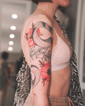 Woman's arm with a snake tattoo intertwined with red poppy flowers. The tattoo covers the upper arm and shoulder, showing intricate detail and shading. Created by our tattoo artists in Canggu, Bali