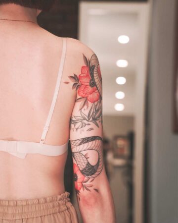 Woman's arm with a snake tattoo intertwined with red poppy flowers. The tattoo covers the upper arm and shoulder, showing intricate detail and shading. Created by our tattoo artists in Canggu, Bali