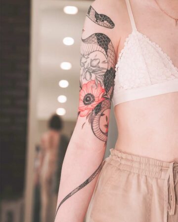 Woman's arm with a snake tattoo intertwined with red poppy flowers. The tattoo covers the upper arm and shoulder, showing intricate detail and shading. Created by our tattoo artists in Canggu, Bali