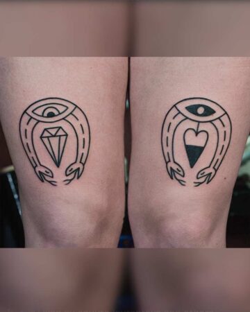 Matching snake tattoos on thighs. Black ink forms two horseshoe shapes, each with snake heads at the bottom, an eye at the top, and a diamond and heart inside. A cool SNAKE TATTOO design. Created by the best tattoo artists in Canggu Bali